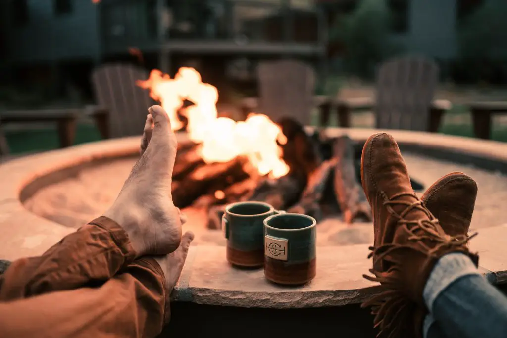Couple with their feet on a fire pit with a fire in it, enjoying hot cocoa.